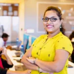 Professional woman smiling in a classroom setting during an online ECCE course in Mumbai, with students attending training sessions in the background.